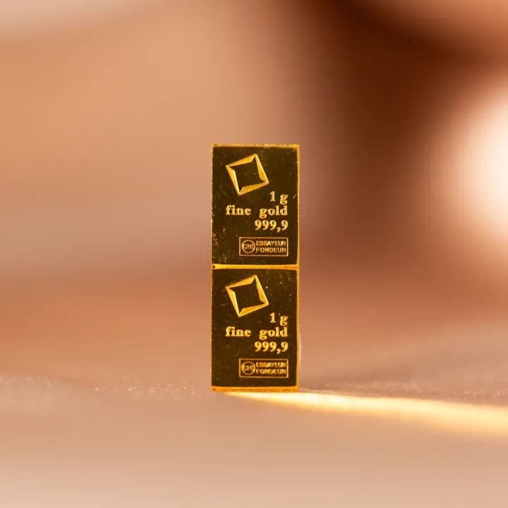 Two stacked 1 gram fine gold bars marked 999.9 and essayeur fondeur, standing on end against a warm blurred background