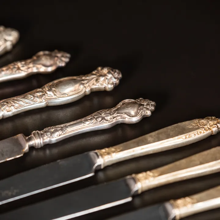 Collection of antique silver-handled knives and ornate flatware pieces laid diagonally on a dark surface