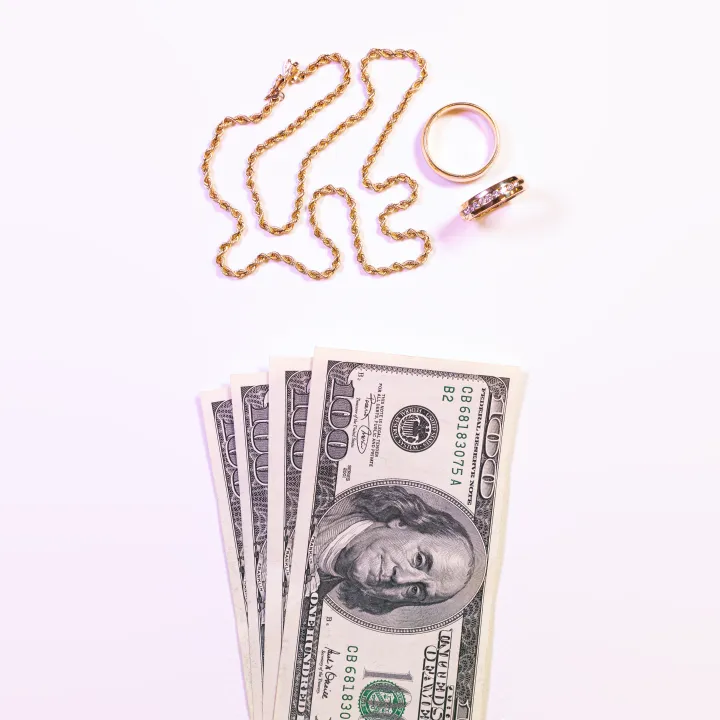 Gold rope chain necklace and two gold rings arranged above a fanned stack of one hundred dollar bills on a light background