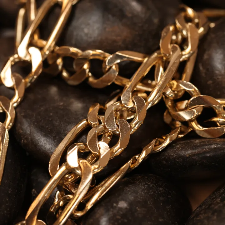 Close-up of several thick gold chain bracelets piled together on dark river stones