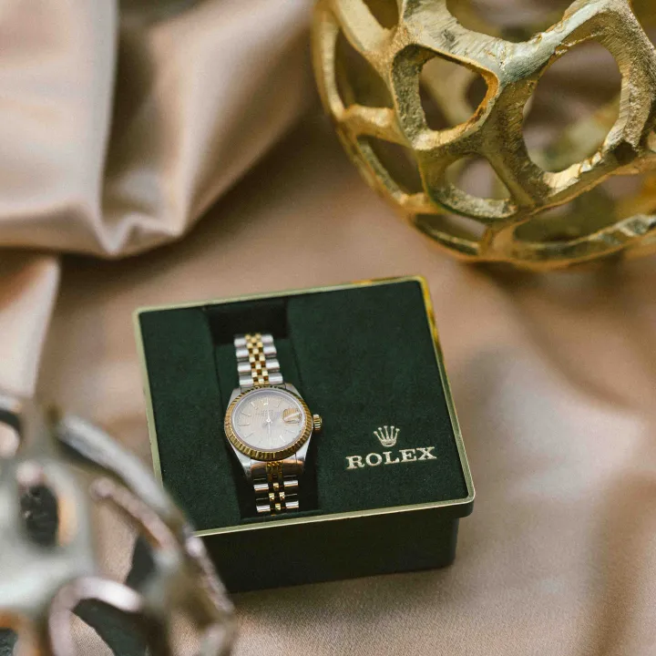 Two-tone Rolex Datejust watch displayed in its black and gold branded box, styled with satin fabric and decorative gold spheres
